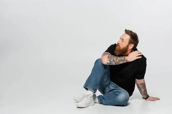Full length of tattooed man in blue jeans and white sneakers sitting and looking away on grey background — Stockfoto