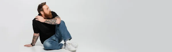 Full length of bearded man in black t-shirt and jeans looking away while sitting on grey background, banner — Stock Photo