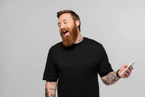 Щасливий татуйований чоловік в чорній футболці тримає смартфон і сміється із закритими очима ізольовано на сірому — стокове фото