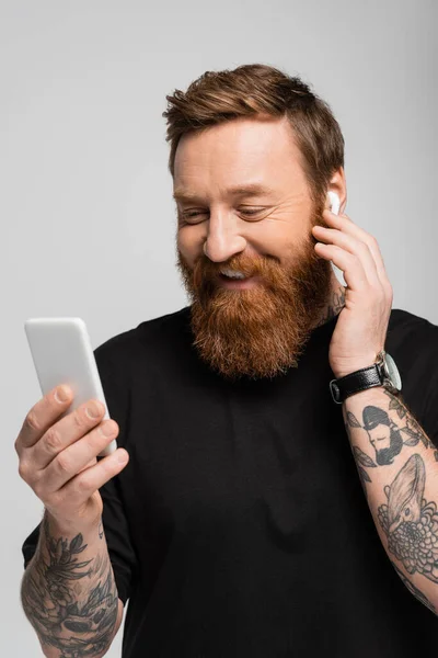 Happy bearded man adjusting wireless earphone while looking at smartphone isolated on grey — Stockfoto