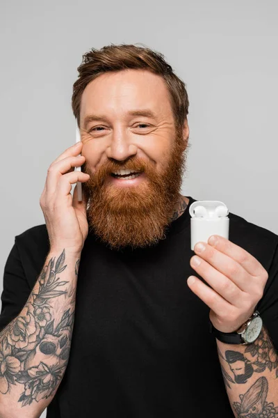 Усміхнений татуйований чоловік тримає футляр з бездротовими навушниками під час розмови на мобільному телефоні ізольовано на сірому — стокове фото
