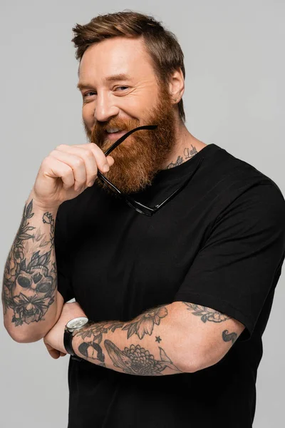 Stylish tattooed man in black t-shirt and wristwatch holding eyeglasses and looking at camera isolated on grey — Stock Photo