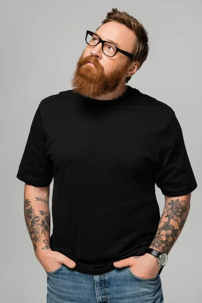 Stylish bearded man in eyeglasses standing with hands in pockets of jeans and looking away isolated on grey — Stock Photo