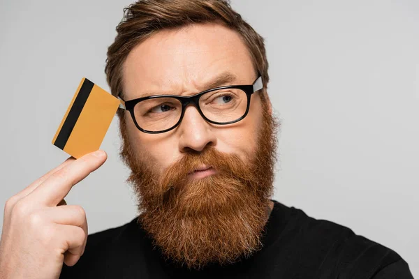 Бородатий чоловік в окулярах тримає кредитну картку біля голови і думає, дивлячись ізольовано на сірому — стокове фото