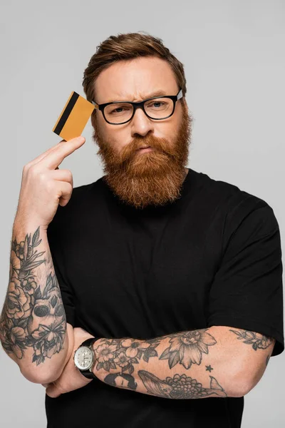 Вдумливий татуйований чоловік в окулярах тримає кредитну картку біля голови, дивлячись на камеру ізольовано на сірому — стокове фото