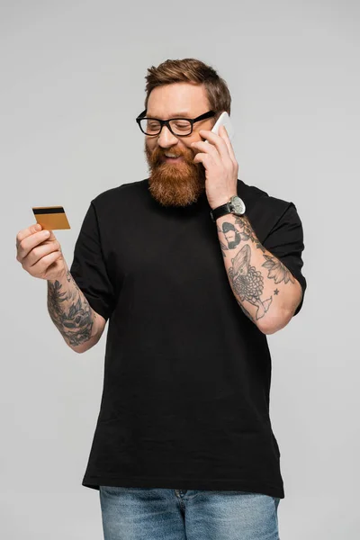 Щасливий татуйований чоловік в окулярах говорить на мобільному телефоні, тримаючи кредитну картку ізольовано на сірому — стокове фото