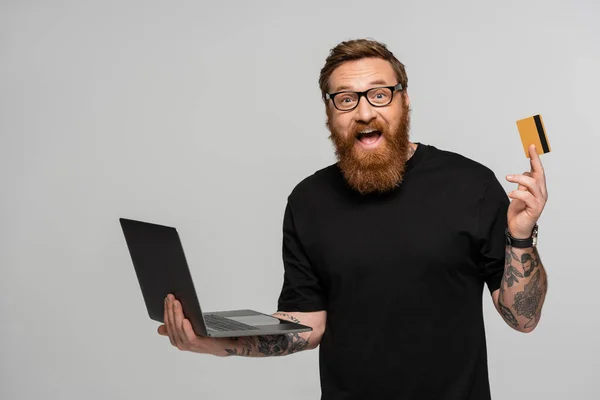 Amazed bearded man in eyeglasses holding laptop and showing credit card isolated on grey — Stock Photo