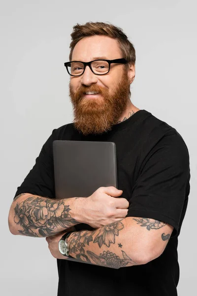 Pleased bearded freelancer in eyeglasses holding laptop and smiling at camera isolated on grey — Photo de stock