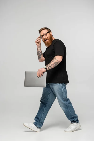 Pleine longueur de lunettes freelance ajustement excité tout en marchant avec ordinateur portable sur fond gris — Photo de stock