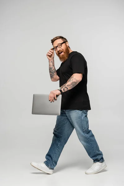 Full length of excited freelancer adjusting eyeglasses while walking with laptop on grey background — Stock Photo