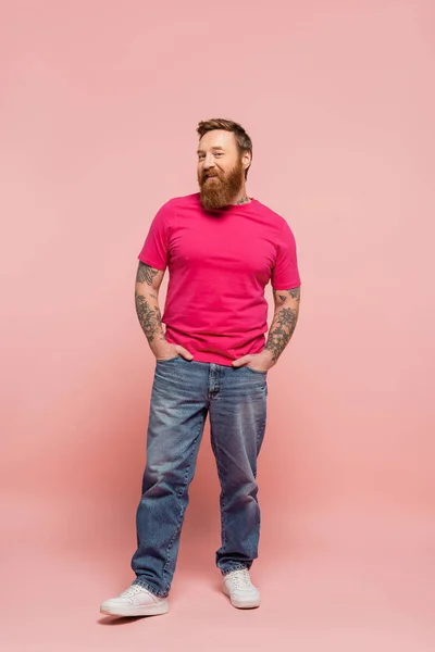 Полная длина счастливый бородатый мужчина в пурпурной футболке и джинсах позирует с руками в карманах на розовом фоне — стоковое фото