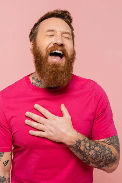 Cheerful bearded man touching chest and laughing with closed eyes isolated on pink — Stock Photo