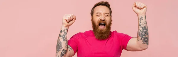 Excited tattooed man with closed eyes shouting and showing win gesture isolated on pink, banner — Stock Photo