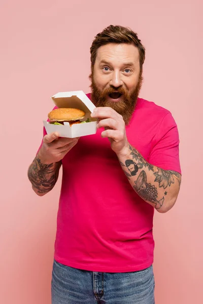 Uomo stupito in magenta t-shirt che mostra scatola di cartone con gustoso hamburger isolato su rosa — Foto stock