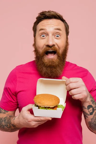 Erstaunt tätowierter Mann in hellem T-Shirt zeigt Kartonpackung mit leckerem Hamburger isoliert auf rosa — Stockfoto