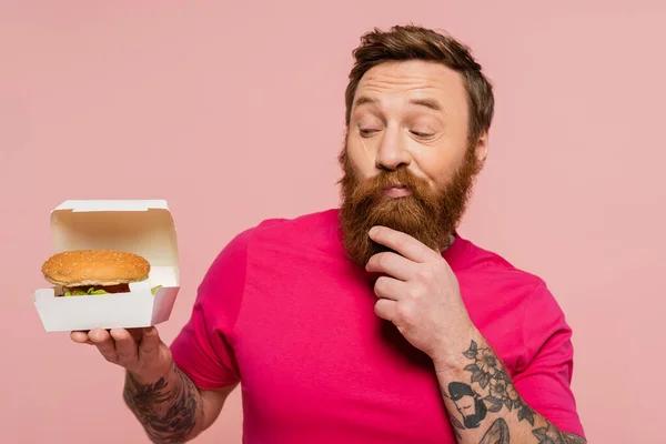 Reflexivo hombre tatuado tocando la barba mientras mira el paquete de cartón con sabrosa hamburguesa aislada en rosa — Stock Photo