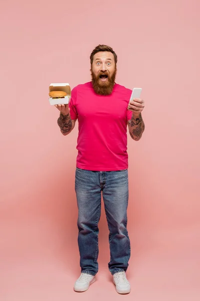 Полный рост изумленного бородатого мужчины, держащего смартфон и гамбургер на розовом фоне — стоковое фото