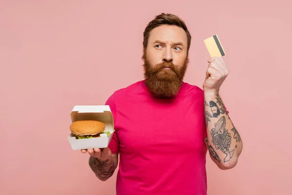 Вдумливий татуйований чоловік тримає кредитну картку та картонну коробку зі смачним бургер, дивлячись ізольовано на рожевому — стокове фото