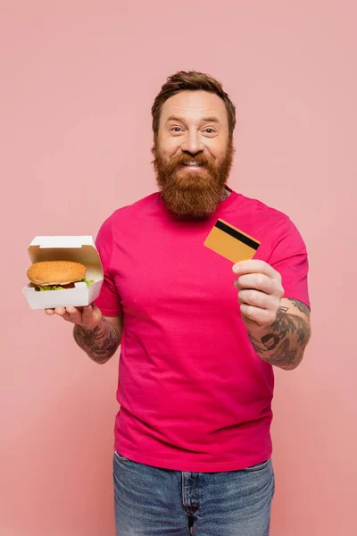 Fröhlicher bärtiger Mann hält Hamburger in der Hand und zeigt Kreditkarte isoliert auf rosa — Stockfoto