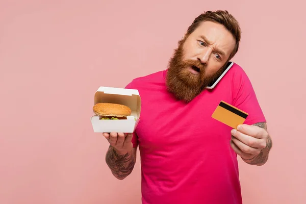 Шокований чоловік дивиться на кредитну картку, тримаючи картонну коробку з бургер і розмовляючи на мобільному телефоні ізольовано на рожевому — стокове фото