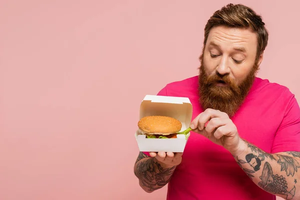 Barbudo hombre tomando papas fritas de paquete de cartón con sabrosa hamburguesa aislado en rosa — Stock Photo