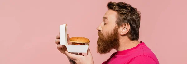 Vista lateral del hombre barbudo oliendo deliciosa hamburguesa aislada en rosa, pancarta - foto de stock