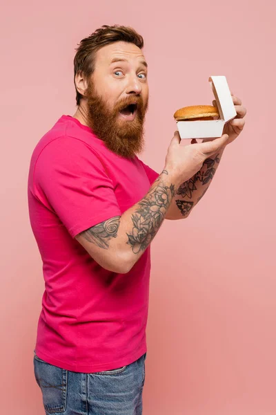 Дивовижний татуйований чоловік з відкритим ротом тримає картонну упаковку з бургер, дивлячись на камеру ізольовано на рожевому — стокове фото