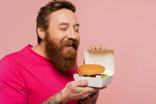 Hombre barbudo complacido con los ojos cerrados sosteniendo paquete de cartón con sabrosa hamburguesa aislada en rosa - foto de stock