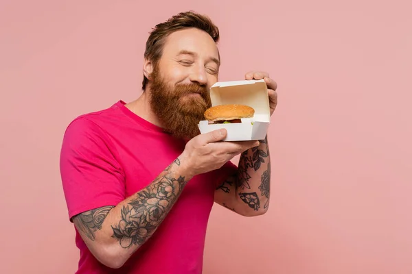 Fröhlich tätowierte Mann mit geschlossenen Augen riecht leckeren Burger in Karton isoliert auf rosa — Stockfoto
