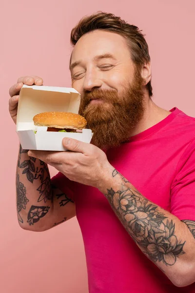 Pleased stylish man with closed eyes smelling tasty burger in carton pack isolated on pink — Stockfoto