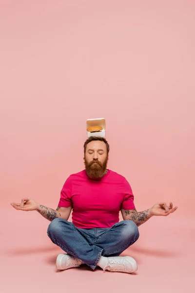 Stylish bearded man with closed eyes and carton box with tasty burger on head meditating in lotus pose on pink background — Foto stock