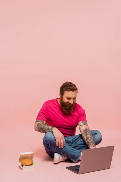 Smiling bearded freelancer working on laptop while sitting with crossed legs near pack with hamburger on pink background — Stock Photo