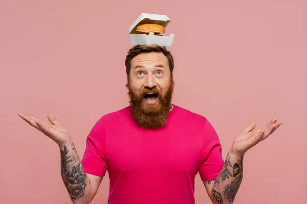 Fröhlicher bärtiger Mann in magenta T-Shirt posiert mit leckerem Burger auf dem Kopf isoliert auf rosa — Stockfoto