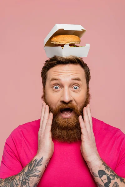Astonished man with hamburger in carton pack on head touching beard and looking at camera isolated on pink — Stockfoto