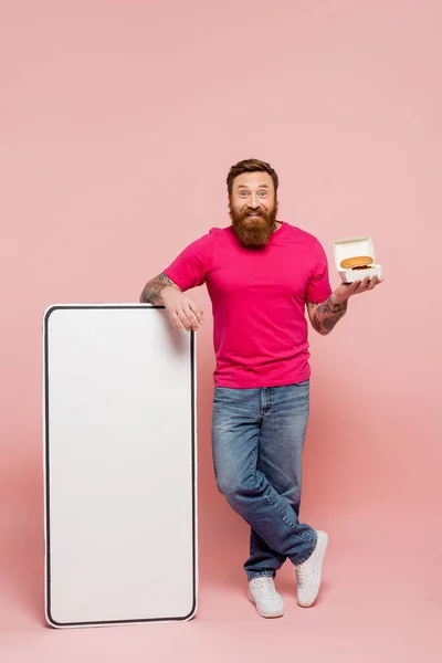 Volle Länge des gut gelaunten Mannes in Jeans steht mit Hamburger in der Nähe große Vorlage des Smartphones auf rosa Hintergrund — Stockfoto