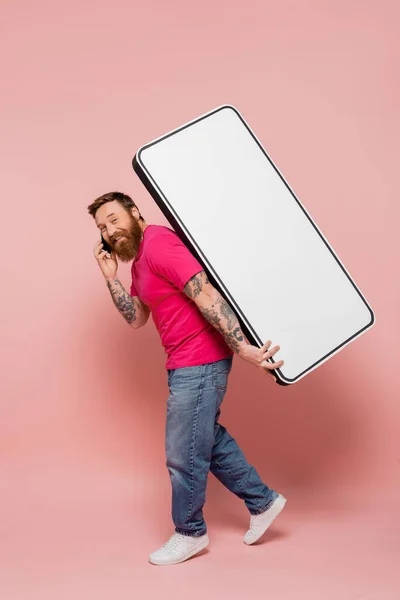 Smiling bearded man carrying huge phone template and talking on smartphone on pink background — Stock Photo