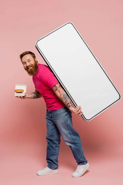 Happy bearded man in magenta t-shirt and jeans carrying hamburger and white template of smartphone on pink background — Stock Photo