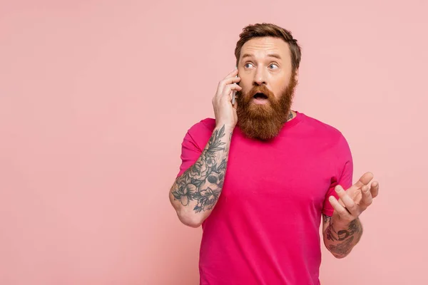 Здивований бородатий чоловік у футболці з пурпуровою сорочкою, дивлячись подалі, розмовляючи на смартфоні ізольовано на рожевому — стокове фото
