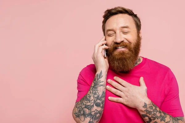 Щасливий татуйований чоловік із закритими очима торкається грудей під час розмови на смартфоні ізольовано на рожевому — стокове фото