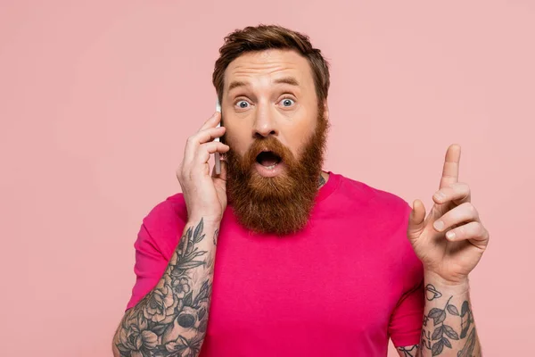 Amazed tattooed man talking on mobile phone and showing idea sign isolated on pink — Stock Photo