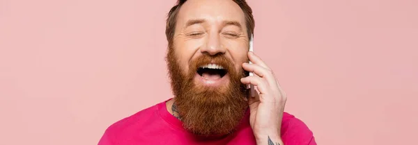 Смеющийся бородач с закрытыми глазами разговаривает на смартфоне изолирован на розовый, баннер — стоковое фото
