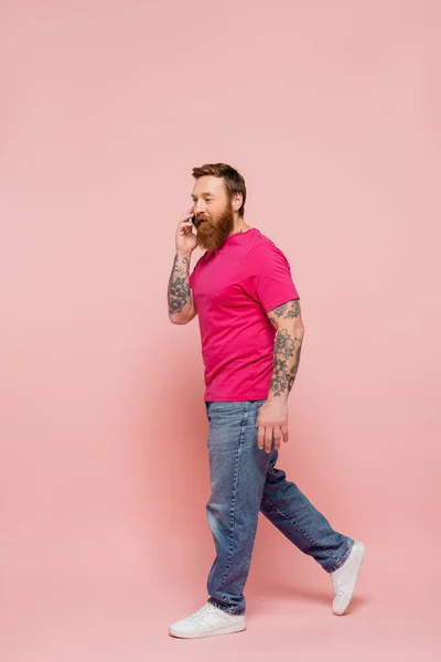 Full length of bearded man in magenta t-shirt and jeans walking during conversation on smartphone on pink background — Photo de stock