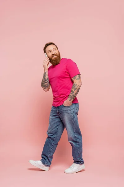 Full length of happy and trendy man talking on cellphone while walking with hand in pocket on pink background — Stock Photo