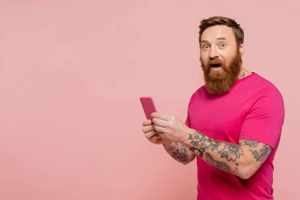 Excited bearded man with open mouth using cellphone and looking at camera isolated on pink — Stock Photo