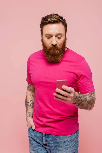 Tattooed man in magenta t-shirt standing with hand in pocket of jeans and using smartphone isolated on pink — Stock Photo