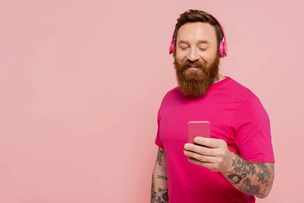 Smiling bearded man in wireless headphones and magenta t-shirt looking at smartphone isolated on pink — Foto stock