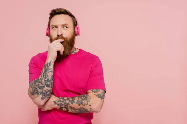 Homme tatoué réfléchi dans des écouteurs sans fil tenant la main près du visage et regardant loin isolé sur rose — Photo de stock