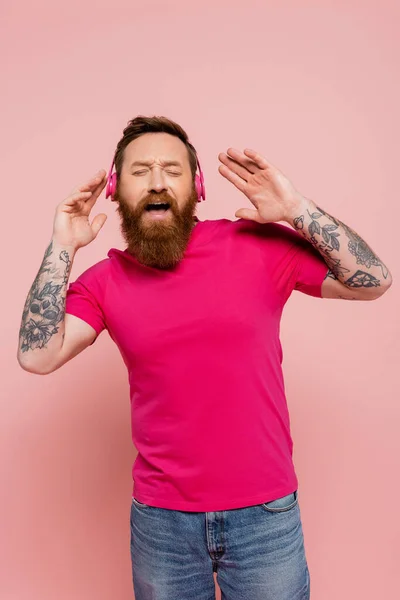 Aufgeregter bärtiger Mann hört Musik in drahtlosen Kopfhörern und singt mit geschlossenen Augen isoliert auf rosa — Stockfoto