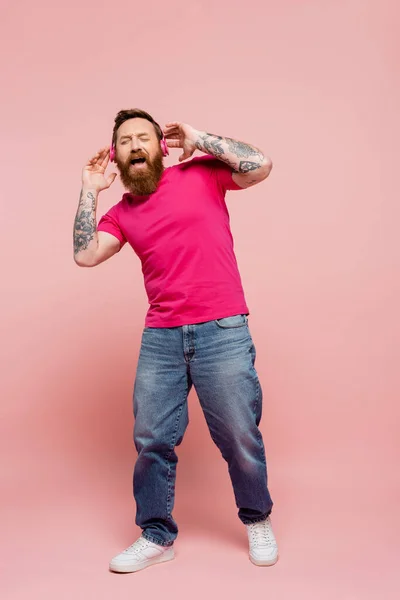Full length of astonished man with closed eyes listening music in wireless headphones and singing on pink background - foto de stock