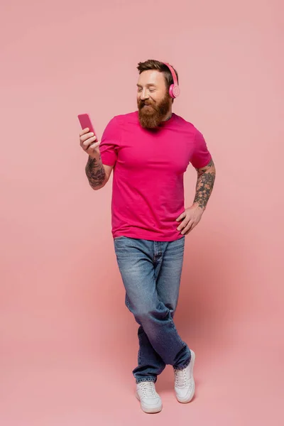 Full length of happy man in wireless headphones holding smartphone while standing with hand on hip on pink background - foto de stock
